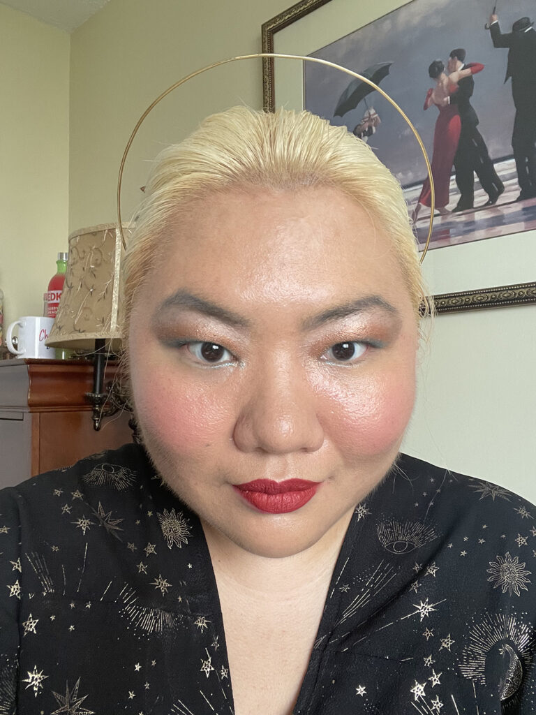 A blonde non-binary woman Amber Ying faces the camera. They are wearing makeup: soft teal blended with bronze, pink blush, and red lipstick. They are also wearing a golden halo above their head.