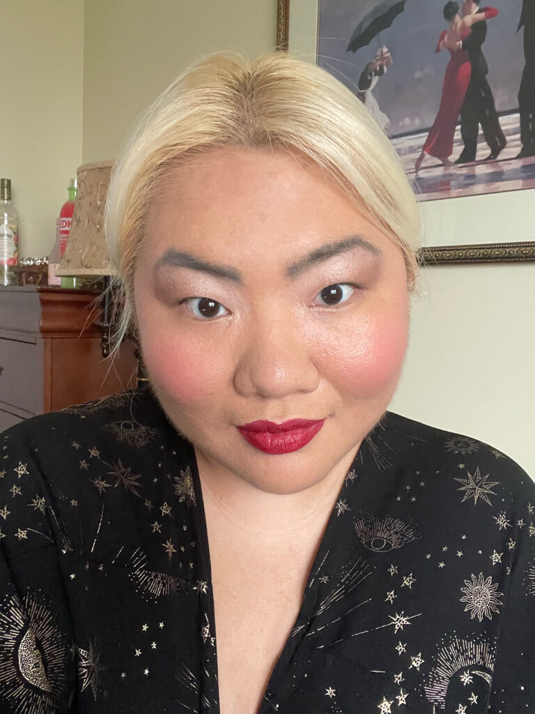 A blonde non-binary woman smiles at the camera. Their hair is parted and pulled back. They are also wearing makeup: a wine red lip, light pink blush, and natural eyeshadow.